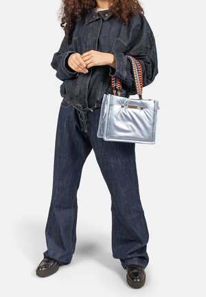 Vrouw draagt een donker denim jack en broek, houdt een metallic zilveren handtas met meerkleurige gevlochten handvatten vast.