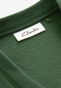 Tmavozelený bavlnený odev s hladkou textúrou a okrúhlym výstrihom. Obsahuje bielu nášivku s výrazne vyšitým logom "Clarks".