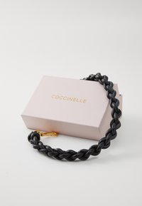 Correa de cuero trenzado negro con un cierre en tono dorado, reposando junto a una caja rosa claro que presenta el texto "COCCINELLE" en dorado.