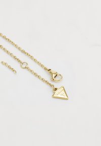 Collana in oro con un pendente triangolare inciso con "GUESS." Presenta un gancio a moschettone e link testurizzati.