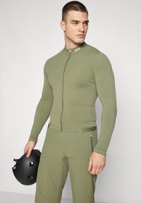 Costume zippé vert olive à manches longues et design ajusté ; texture lisse. Casque noir tenu à la main, avec des trous de ventilation.