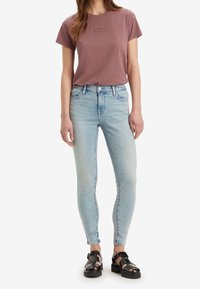 Jeans ajustados de color azul claro, combinados con una camiseta rosa empolvado que presenta un logo oscuro. Sandalias negras con tiras completan el conjunto.