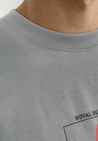 Pilkos medvilnės marškinėliai su pailgu apykakle, su grafiniu piešiniu ir juodu tekstu "ROYAL DEPARTMENT". Artimas medžiagos tekstūros vaizdas.