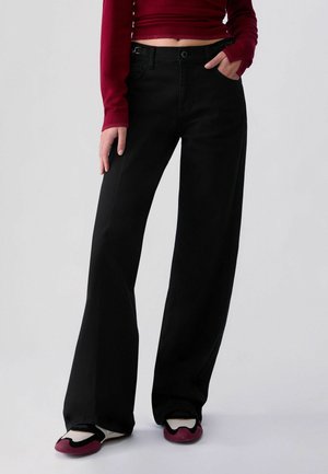 Personne portant un pantalon noir taille haute à jambes larges, un top court bordeaux à manches longues et des baskets noires et bordeaux, debout avec une main dans la poche.
