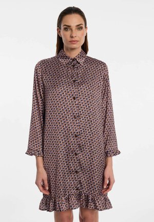 Woman wearing a long-sleeve, buttoned dress with a geometric pattern and ruffled cuffs and hem, standing against a plain light background.