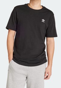 Svart bomulls t-shirt med kort ärm, rund halsringning och vit Adidas-logotyp på vänster bröst. Bärs med gråa sweatpants.