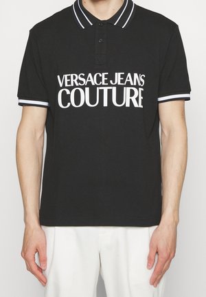 Polo noir à manches courtes avec des rayures blanches sur le col et les manches, avec un grand texte blanc "VERSACE JEANS COUTURE" sur le devant.