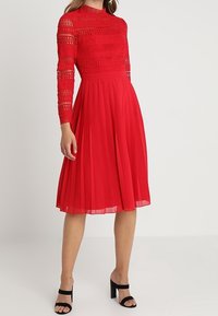 Červené šaty s plisovanou sukní, dlouhými krajkovými rukávy a vysokým výstřihem. Hladká měkká textura látky s propracovanými krajkovými detaily.