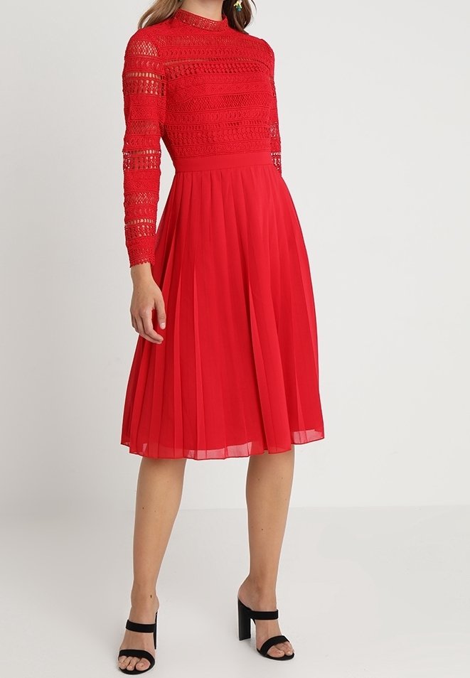 Červené šaty s plisovanou sukní, dlouhými krajkovými rukávy a vysokým výstřihem. Hladká měkká textura látky s propracovanými krajkovými detaily.