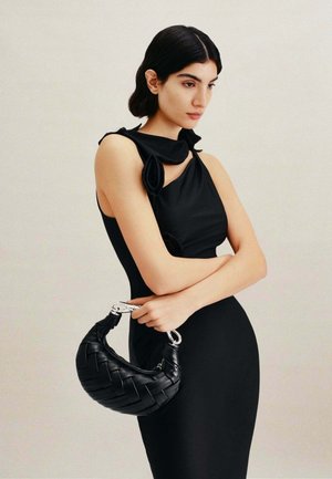 Femme aux cheveux foncés portant une robe noire sans manches tenant un sac à main noir tressé avec une poignée argentée sur un fond neutre.