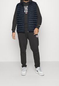 Granatowa kamizelka puchowa z pikowaną teksturą na czarnej bluzie z kapturem i dresach. Białe sneakersy dopełniają zestaw, tworząc sportowy i swobodny wygląd.