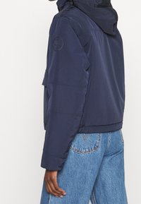 Person som bär en marinblå huvtröja med en logotyp på ärmen och ljusblå denimjeans, visad från mitten av ryggen till mitten av låret.