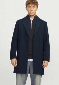 Abrigo de lana azul oscuro con cuello muesca, mangas largas, cuatro botones y bolsillos laterales, superpuesto sobre una camiseta negra con cremallera y jeans claros.