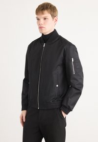 Black bomber jacket with elastic cuffs and hem, silver zip closure, side pockets, and zippered pocket on left sleeve. Smooth texture.
