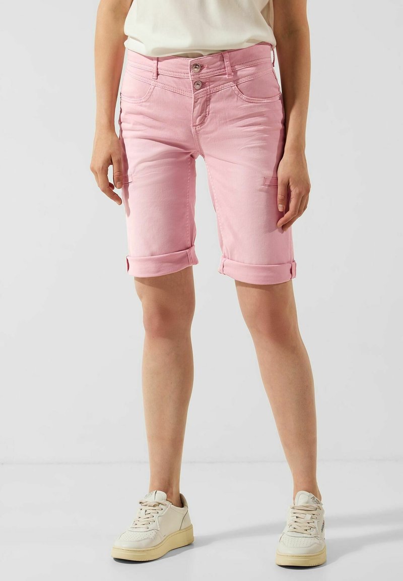 Personne portant un short rose remonté jusqu'aux genoux et des baskets blanches, debout devant un fond clair uni.