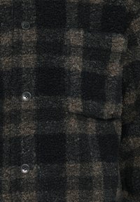 Cămașă din flanel în nuanțe închise de negru și maro, având un model cu pătrățele și nasturi argintii. Materialul are un finish texturat și pufos.