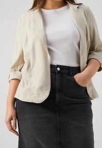 Jasnobeżowy lniany blazer z podwiniętymi rękawami, dwoma kieszeniami, noszony na białej prążkowanej koszulce, w zestawieniu z czarną dżinsową spódnicą o wysokim stanie.