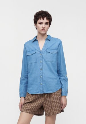 Jeune personne aux cheveux courts et bouclés, portant une chemise bleu clair à boutons et un short à carreaux, debout devant un fond blanc uni.