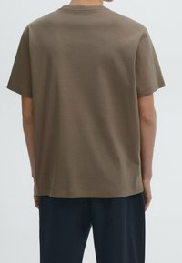 Person wearing a plain brown short-sleeve t-shirt and dark blue pants, shown from the back against a plain background.