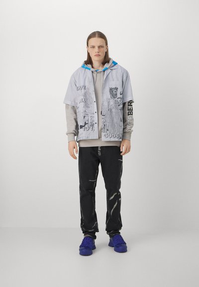 Gestreiftes Kurzarmhemd, das über einen grauen Hoodie getragen wird, kombiniert mit schwarzen Hosen mit weißen Streifen und blauen Turnschuhen.