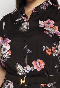 Mustapohjainen kukkamekko, jossa on kaulus, lyhyet hihat ja kangasvyö. Mekossa on värikkäitä kukkakuvioita ja kultainen solkisomiste.