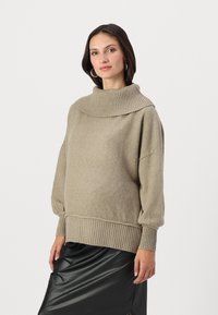 Pull en tricot beige doux avec un col montant à côtes, des épaules tombantes, et des poignets et un ourlet à côtes. Assorti à une jupe noire brillante.