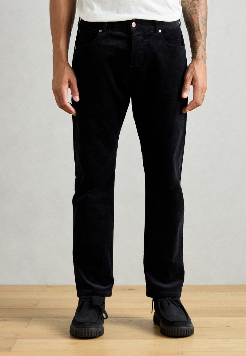 Pantalons en velours côtelé noirs avec une coupe droite, des détails de poches, associés à des chaussures noires sur un sol en bois.