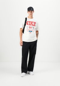 White t-shirt with red and blue graphic design, black cargo pants, and a black cap. White sneakers complete the outfit.