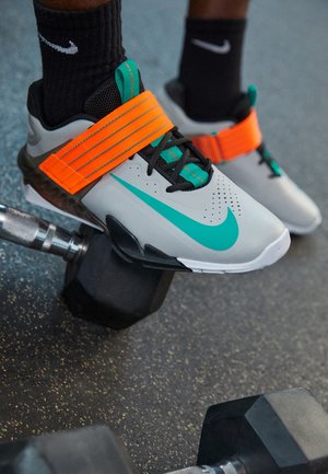Gros plan sur des chaussures d'haltérophilie Nike grises et turquoise avec des sangles orange aux pieds portant des chaussettes noires Nike, positionnées près d'haltères noirs sur le sol de la salle de sport.