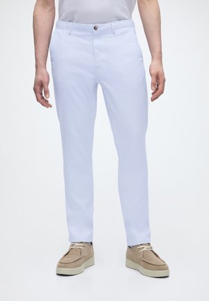 Uomo con pantaloni bianchi slim fit e scarpe beige in suede con lacci su sfondo bianco semplice.