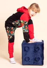 Czerwona i czarna bluza z kapturem w połączeniu z kwiatowymi spodniami; materiał jest miękki i elastyczny. Białe trampki na rzepy. Niebieska pluszowa kostka jako rekwizyt.