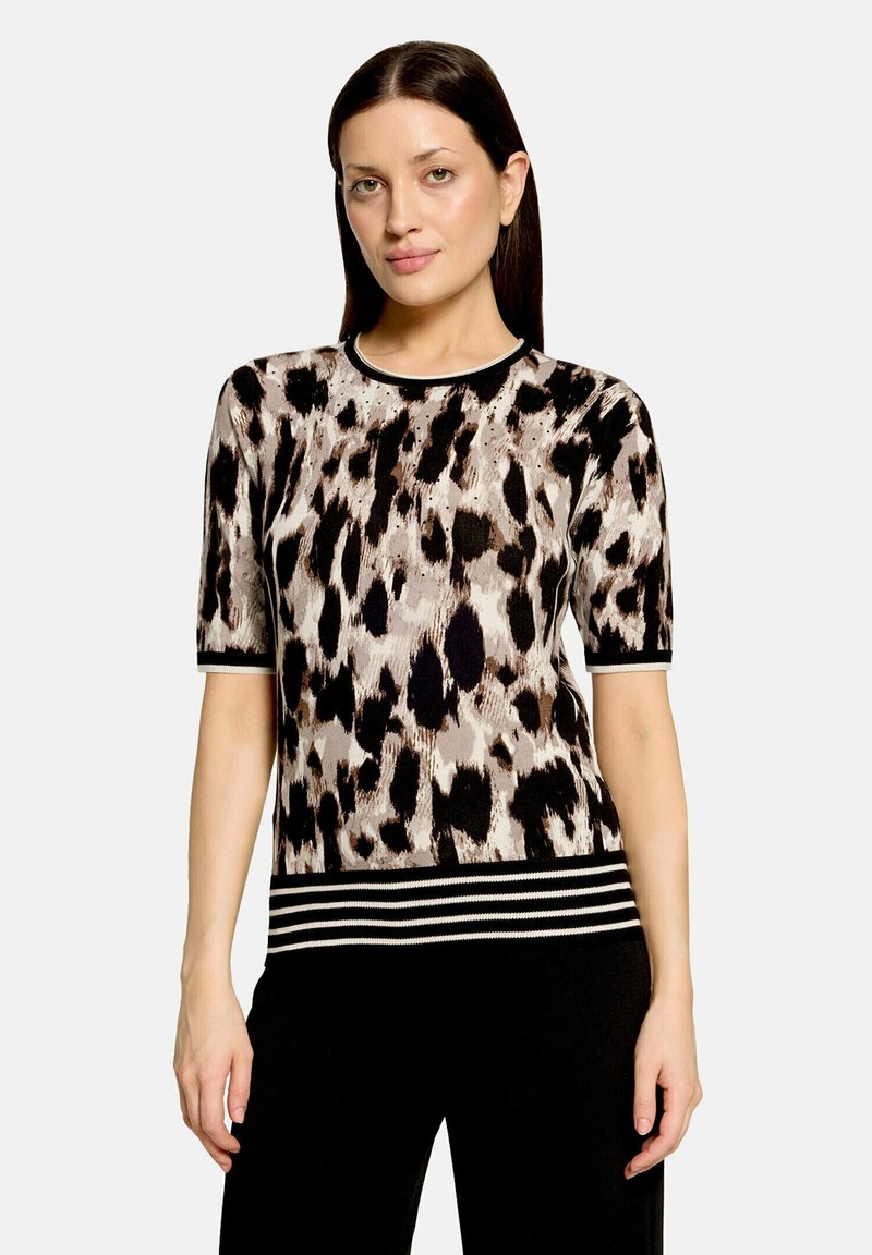 Femme aux cheveux raides et foncés portant un haut à manches courtes imprimé animal avec un ourlet rayé noir et blanc et un pantalon noir sur un fond uni.