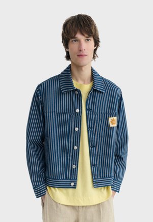 Jeune homme portant une veste rayée verticale bleue et blanche par-dessus une chemise jaune et un pantalon beige, debout devant un fond gris uni.