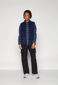 Mornarski telovnik iz flisa preko modre karo srajce, kombiniran s črnimi cargo hlačami in belimi supergami. Model stoji pred enobarvnim ozadjem.