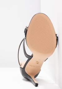 Musta korkea kantapää sandaalit nilkkahihnalla asetettuna pohja ylöspäin, osoittaen sileää vaaleanruskeaa pohjaa, jossa on teksturoitu ote ja pieni kultainen brändiembleemi.