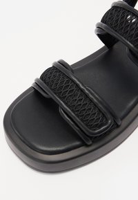 Mustat sandaalit, joissa on sileä, muotoiltu pohja ja tekstuurilla kudotut hihnat. Varustettu kahdella säädettävällä hihnalla varman istuvuuden takaamiseksi.