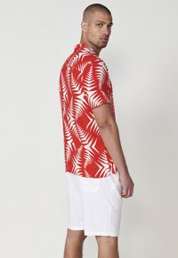 Rotes kurzärmeliges Hemd mit einem weißen tropischen Blättermuster, kombiniert mit weißen Shorts. Das Hemd hat einen geraden Saum und eine lockere Passform.