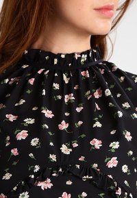 Black fabric blouse with a gathered neck, featuring a floral pattern in pink and white, with ruffled accents at the chest and waist areas.
