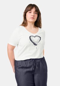 Weißes Kurzarm-T-Shirt mit einem Grafikdesign eines marineblauen Herzens mit Punktmustern, kombiniert mit dunklen, lässigen Hosen mit elastischem Bund.