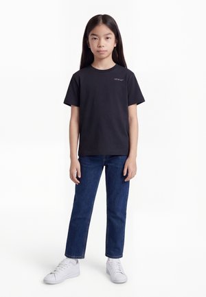 Νεαρό άτομο με μακριά σκουρόχρωμα μαλλιά που φοράει μαύρο T-shirt Off-White, μπλε τζιν και λευκά αθλητικά παπούτσια, στέκεται μπροστά σε λευκό φόντο.