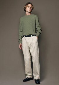 Pull en rib à col olive, pantalon plissé écru, ceinture sombre, mocassins noirs avec accents dorés, design minimaliste sur fond neutre.