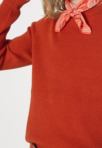 Personne portant un pull orange côtelé et un bandana orange à motifs noué autour du cou, sur un fond uni.