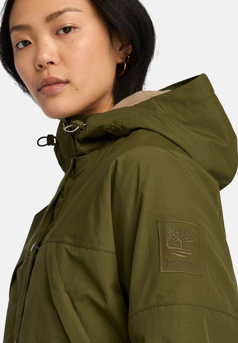 Giacca impermeabile verde oliva con cappuccio, dotata di una fodera in beige, collo regolabile e un'etichetta con il logo Timberland sulla manica.