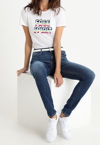Camiseta blanca con "FREE SPIRIT" en una tipografía retro colorida. Combinada con unos jeans ajustados de color azul oscuro y zapatillas blancas, sentado sobre un cubo blanco.