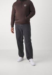 Uomo che indossa un hoodie marrone scuro con una grafica del sole, jeans neri e sneaker bianche con uno swoosh dorato, appoggiato su uno sfondo semplice.