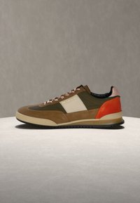 Χαμηλό σνίκερ με καφέ, ελιά πράσινο, μπεζ και πορτοκαλί πάνελ, κρέμ σόλα και ροζ-λευκά κορδόνια σε ουδέτερο φόντο.