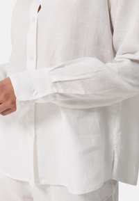 Camicia bianca in lino a maniche lunghe con un davanti a bottoni, sottile trama, spacchi laterali e polsini con bottoni rotondi e trasparenti.