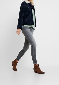 Žena jde oblečená v tmavě modré kordové bundě s bílým límcem, světle zeleném svetru, šedých slim fit džínech a hnědých kotní�čkových botách.