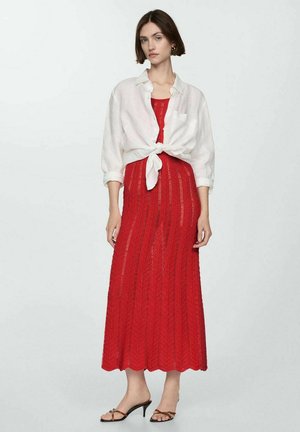 Woman wearing a red knitted sleeveless midi dress with a white button-up shirt tied at the waist and black heeled sandals.