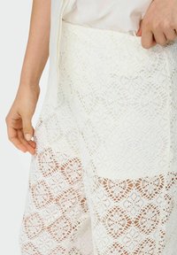 Pantaloncini bianchi in pizzo con motivi floreali intricati, una finitura testurizzata e una cintura che viene regolata con una mano.